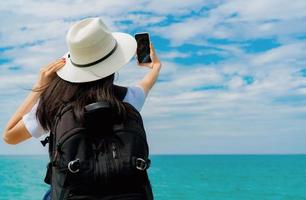 la giovane donna asiatica con zaino e sacco a pelo indossa il cappello usa lo smartphone prendendo selfie al molo. vacanze estive sulla spiaggia del paradiso tropicale. felice hipster ragazza viaggiare in vacanza. la donna si diverte e si rilassa la vita. vibrazioni estive. foto