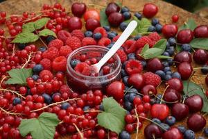 cucchiaio bianco con marmellata di frutti di bosco nel bicchiere. mirtillo, lampone, ciliegia e ribes sul tavolo di legno in giardino. foto