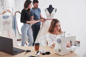 la ragazza e l'uomo misurano il vestito. stilista femminile lavora sui nuovi vestiti in officina con un gruppo di persone dietro foto