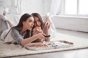 tempo libero divertente. sorelle che mangiano pizza quando guardano la tv sdraiate sul pavimento di una bella camera da letto durante il giorno foto