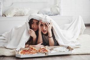 è così spaventoso. coperta calda. sorelle che mangiano pizza quando guardano la tv sdraiate sul pavimento di una bella camera da letto durante il giorno foto