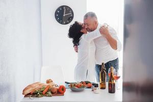 tempo per abbracciarsi e ballare. l'uomo anziano e sua moglie in camicia bianca hanno una cena romantica in cucina foto