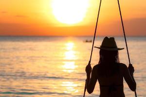 silhouette donna indossare bikini e cappello di paglia oscillare le altalene in spiaggia durante le vacanze estive al tramonto. ragazza che si diverte e si rilassa in vacanza. vibrazioni estive. donna che guarda il bel cielo al tramonto. foto