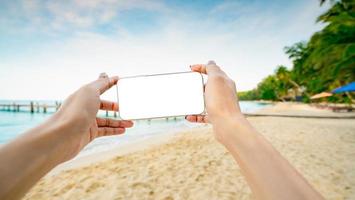 mani della donna che tengono smartphone con display vuoto sulla spiaggia di sabbia dell'isola tropicale. donna che scatta una foto della spiaggia paradisiaca durante le vacanze estive. donna asiatica adulta godendo e rilassante sulla spiaggia.