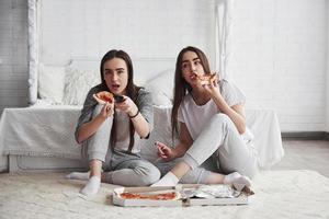 il cibo è sempre più delizioso quando si guardano i film. sorelle che mangiano pizza davanti alla tv mentre si siedono sul pavimento della bella camera da letto durante il giorno foto