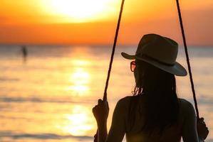 silhouette donna indossare bikini e cappello di paglia oscillare le altalene in spiaggia durante le vacanze estive al tramonto. ragazza in costume da bagno si siede sulle altalene e guarda il bel tramonto. vibrazioni estive. la donna viaggia da sola. foto