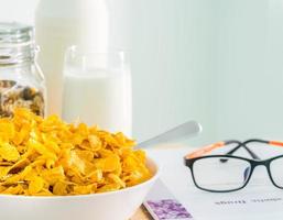 ciotola di cereali con cucchiaio e un bicchiere di latte messo su un tavolo di legno vicino al libro di testo e agli occhiali del diabete. colazione a base di calcio. cornflakes e concetto di latte foto