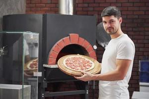 lavoratore amichevole. fornaio in camicia bianca con pizza pronta per essere messa in forno a cuocere foto