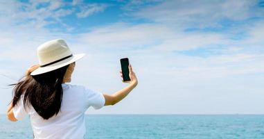 la giovane donna asiatica indossa il cappello in stile casual usa lo smartphone prendendo selfie al molo. vacanze estive sulla spiaggia del paradiso tropicale. ragazza felice viaggia in vacanza. la donna si diverte e si rilassa la vita. vibrazioni estive. foto