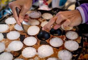 la pasta tostata al mortaio o kanom krok è un dolce tradizionale tailandese. mano della donna che rimuove kanom krok dalla stufa a cucchiaio. cibo di strada in Thailandia. dessert tailandese a base di latte di cocco e farina. foto