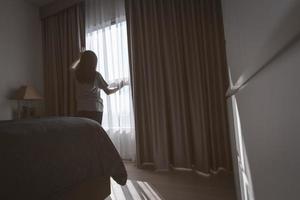 vista posteriore della donna che tiene la tenda e guarda fuori dalla finestra di vetro. donna depressa dal blocco e dalla quarantena dalla pandemia di coronavirus. salute mentale e demenza. donna premurosa in camera da letto. foto