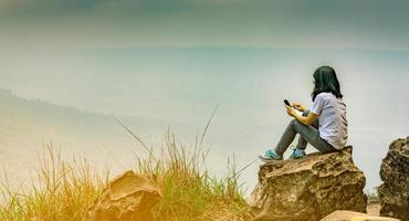 la giovane donna asiatica si siede sulla roccia sulla scogliera e usa il messaggio dello smartphone ai suoi amici mentre è in vacanza al parco nazionale. concetto di viaggio e comunicazione avventurosa foto