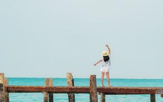 vista posteriore di felice giovane donna asiatica in stile casual moda e cappello di paglia rilassarsi e godersi le vacanze in spiaggia paradiso tropicale. ragazza stand al molo di legno del resort durante le vacanze estive. vibrazioni estive. foto