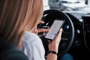 guardare la mappa tramite smartphone. bella ragazza bionda seduta nella nuova auto con interni neri moderni foto