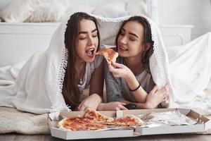 sembra delizioso. coperta calda. sorelle che mangiano pizza quando guardano la tv sdraiate sul pavimento di una bella camera da letto durante il giorno foto