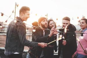 ragazzo con il cappello e la barba si chiede se la fiamma può essere più grande se soffia nel telefono. giocando con le stelle filanti sul tetto. gruppo di giovani bellissimi amici foto