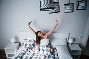 telefono in mano. bella giovane donna sdraiata sul letto al mattino nella sua stanza foto