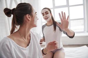 sorridente durante il processo. concezione della cura della pelle utilizzando una maschera bianca sul viso. due sorelle hanno il fine settimana in camera da letto foto