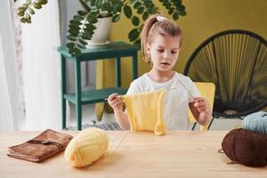 concentrato sul processo. il bambino sta lavorando a maglia a casa. la bambina sveglia che si siede vicino al tavolo di legno sta imparando alcune cose nuove foto