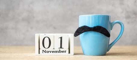 calendario del 1 novembre, tazza di caffè blu o tazza da tè e decorazioni di baffi neri sul tavolo. festa degli uomini, felice festa del papà e ciao concetto di novembre foto