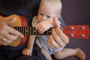 il padre barbuto con i bambini gioca sull'ukulele al coperto foto
