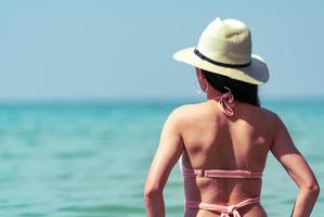 il punto di vista posteriore della giovane donna asiatica sexy indossa bikini rosa, cappello di paglia e occhiali da sole che si rilassano e si godono le vacanze sulla spiaggia del paradiso tropicale. ragazza alla moda per le vacanze estive. modella sexy di bellezza. vibrazioni estive. foto