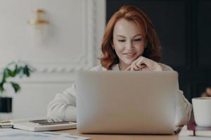 donna d'affari dai capelli rossi esperta legge la pubblicazione su un moderno computer portatile, si concentra sullo schermo con un sorriso piacevole, fa ricerche per la pianificazione dell'avvio, controlla la casella di posta, lavora da casa foto