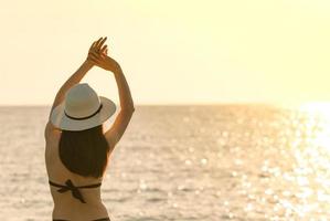 il punto di vista posteriore della giovane donna asiatica felice in costume da bagno nero e cappello di paglia si rilassa e si gode la vacanza sulla spiaggia del paradiso tropicale al tramonto. ragazza alla moda per le vacanze estive. modella sexy di bellezza. stile di vita elegante foto