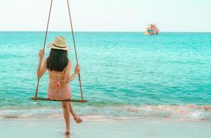 donna asiatica indossare costumi da bagno e cappello oscillare le altalene sulla spiaggia di sabbia e guardare il bellissimo paradiso tropicale mare e cielo in una giornata di sole. vacanze estive. vibrazioni estive. ragazza che si diverte e si rilassa in vacanza foto