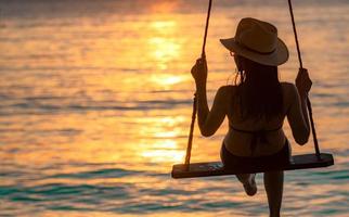 silhouette donna indossare bikini e cappello di paglia oscillare le altalene in spiaggia durante le vacanze estive al tramonto. ragazza che si diverte e si rilassa in vacanza. vibrazioni estive. donna che guarda il bel cielo al tramonto. foto