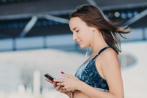 inquadratura laterale di una bella donna sportiva con i capelli scuri, vestita con abiti casual, tiene in mano uno smartphone moderno, controlla le notifiche, ascolta musica con gli auricolari, gode di una connessione Internet ad alta velocità foto