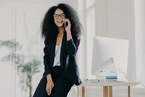 immagine di una donna d'affari soddisfatta dalla pelle scura si appoggia al tavolo bianco con il computer, fa una pausa dopo il lavoro, posa nel suo armadietto, parla tramite cellulare, indossa occhiali e abito elegante, si consulta con il partner foto