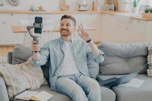 giovane positivo che mostra il segno di pace durante la videochiamata mentre è seduto sul divano foto