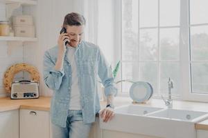 bell'uomo d'affari barbuto impegnato in abiti comodi e casual in piedi in cucina con il telefono cellulare foto