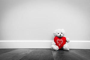 simpatico orsacchiotto con grande cuore di peluche rosso. seduto sul pavimento di legno contro il muro bianco. foto