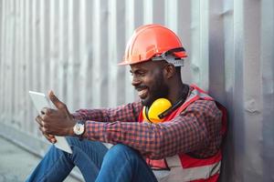 primo piano ingegnere afroamericano maschio sorridente felice che indossa giubbotto di sicurezza e casco, seduto e utilizzando il telefono cellulare nel cantiere di container di carico logistico. foto