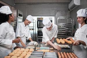 team di gourmet professionisti multirazziali, quattro chef in divise da cuoco bianche e grembiuli impastano pasta e farina, preparano pane e cibi da forno, cuociono in forno nella cucina del ristorante in acciaio inossidabile. foto