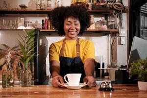 La barista afroamericana offre una tazza di caffè al cliente con un sorriso allegro, un servizio felice lavora in un bar ristorante informale, giovane imprenditore di avvio di piccole imprese. foto