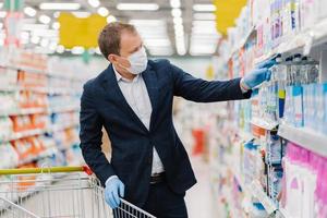 immagine del consumatore uomo acquista detersivo nel centro commerciale, prende una bottiglia di gel detergente nel carrello, sceglie prodotti per la casa, si preoccupa dell'igiene e della protezione durante la situazione pandemica, diffusione del virus foto