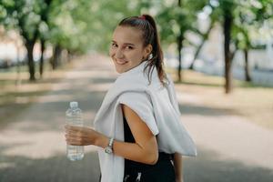 vista posteriore di una donna sportiva allegra e felice con coda di cavallo rimane idratata dopo l'allenamento cardio nel parco tiene una bottiglia di acqua fresca si prende una pausa dopo un faticoso jogging mattutino va regolarmente per lo sport foto