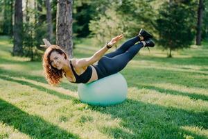 sorridente felice bruna donna con aspetto europeo saldi su palla fitness allunga le braccia vestite in alto e leggings pone all'aperto nel parco estivo facendo pilates in natura durante la giornata di sole foto