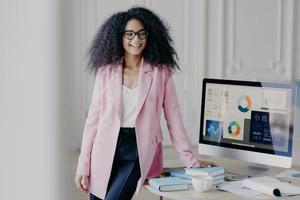 mezza lunghezza di allegra donna afroamericana con i capelli ricci, vestita con un abito formale elegante, posa vicino al tavolo, schermo del computer con grafica, si prepara per la presentazione davanti all'auditorium foto