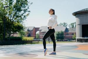 foto a figura intera di atletica donna sportiva si riposa dopo l'aerobica pilates allenamento gode di attività sportive all'aperto indossa felpa bianca leggings e scarpe da ginnastica ascolta musica tiene le mani in vita