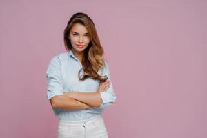 mezza lunghezza di una modella femminile dall'aspetto piacevole indossa camicia e jeans bianchi, tiene le braccia conserte guarda con sicurezza alla telecamera, ha i capelli lunghi, posa su sfondo viola, spazio vuoto per la promozione foto