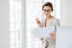 foto di una giovane donna soddisfatta con i capelli scuri vestita in tailleur, concentrata sulle carte, lavora in ufficio, tiene in mano un cellulare moderno, indossa occhiali ottici per una buona visione, ha un'espressione soddisfatta