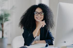 studentessa internazionale positiva con i capelli crespi, indossa occhiali trasparenti, tiene la penna in mano, fa i conti, si siede davanti al grande schermo del computer, vestita con un abito nero elegante. foto