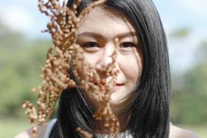 bella donna asiatica in abito bianco rilassarsi e sorridere con steli di legno secco nel parco naturale. la giovane ragazza tailandese si diverte in vacanza con la luce del sole in giardino. foto