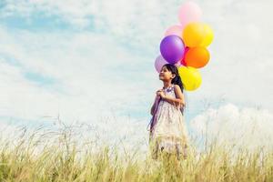 bambina sveglia che tiene palloncini colorati nel prato contro il cielo blu e nuvole, allargando le mani. foto