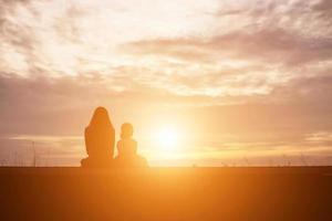 una silhouette di una bambina felice le braccia della sua amorevole madre per un abbraccio, davanti al tramonto nel cielo in una giornata estiva. foto