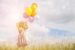 bambina sveglia che tiene palloncini colorati nel prato contro il cielo blu e nuvole, allargando le mani. foto